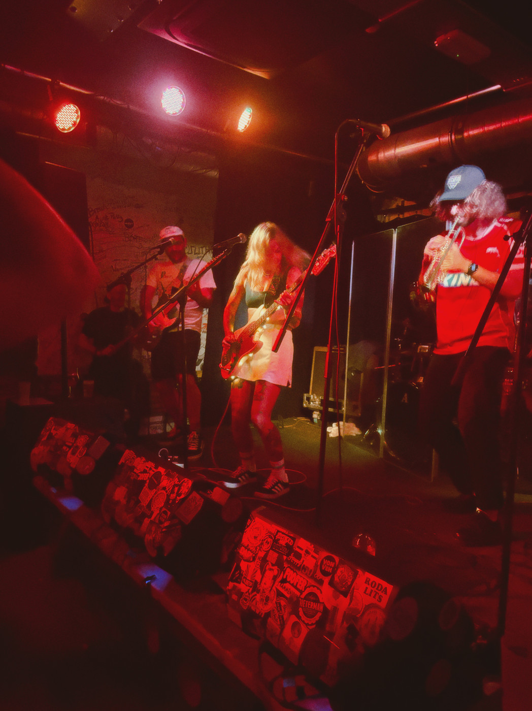 A live band performing on a small club stage bathed in red lighting. Three musicians are visible: on the left, a guitarist wearing a cap and light shirt; in the center, a female guitarist with long blonde hair wearing a light dress; on the right, a saxophonist in a red shirt and blue cap. They stand in front of microphones with gear and amps around, and a row of posters or flyers runs along the edge of the stage. The audience is in the dark foreground.