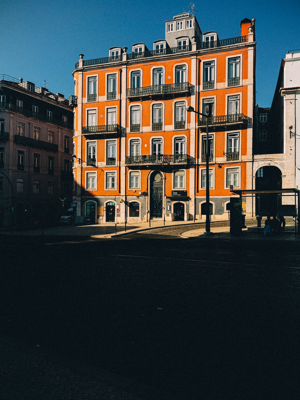 Bright orange historic building with multiple balconies and large windows, partially in shadow, on a sunny street corner with a clear blue sky.