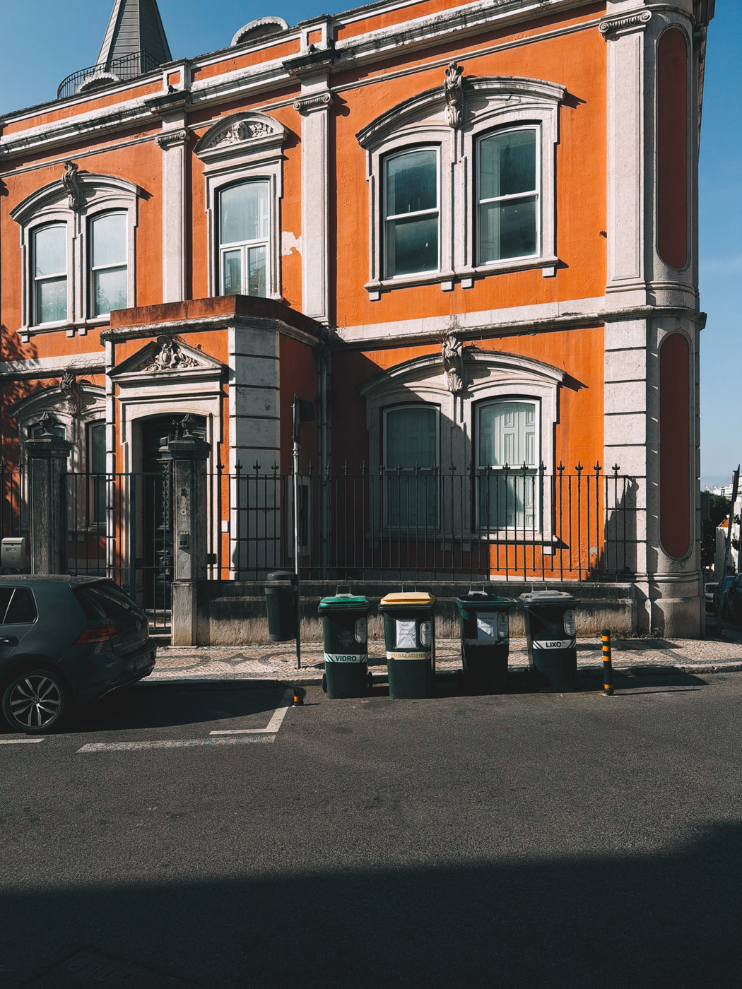 Historic orange building with white trim and arched windows, a black iron fence, parked car, and four green and yellow recycling bins on a sunny street.