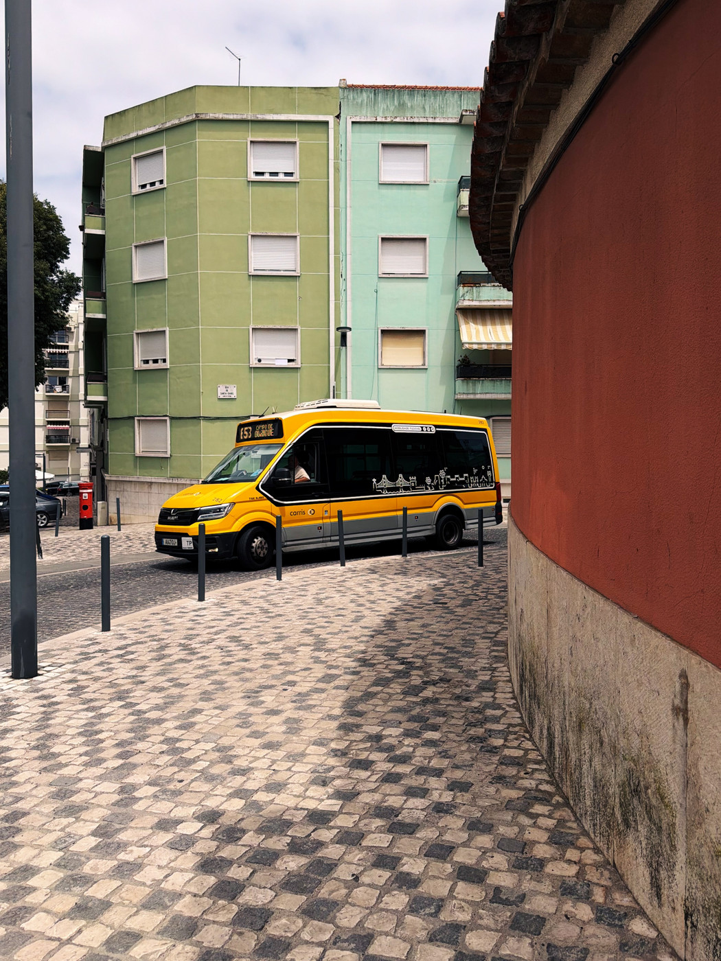 A yellow city minibus marked “153” drives along a cobblestone street lined with pastel green apartment buildings and a curved red wall under a partly cloudy sky.