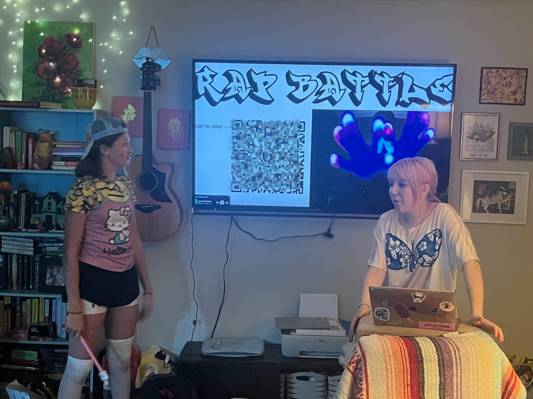 In a cozy colorful cluttered living room two teen girls stand in front of a large wall mounted tv that has the words “rap battle” above a QR code