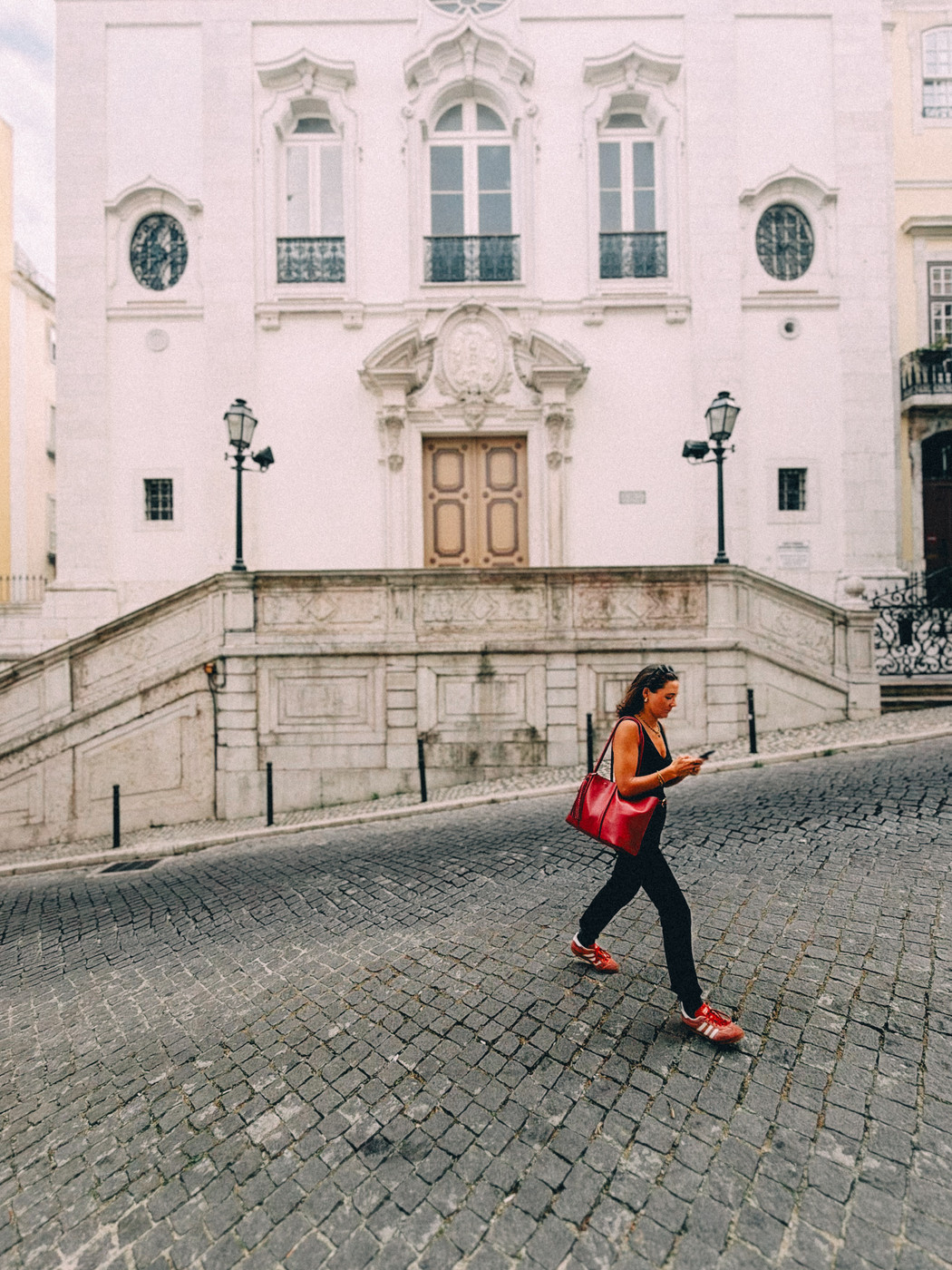 Woman with a red bag and red shoes walking on a cobblestone street in front of a historic church with ornate windows and a central staircase.