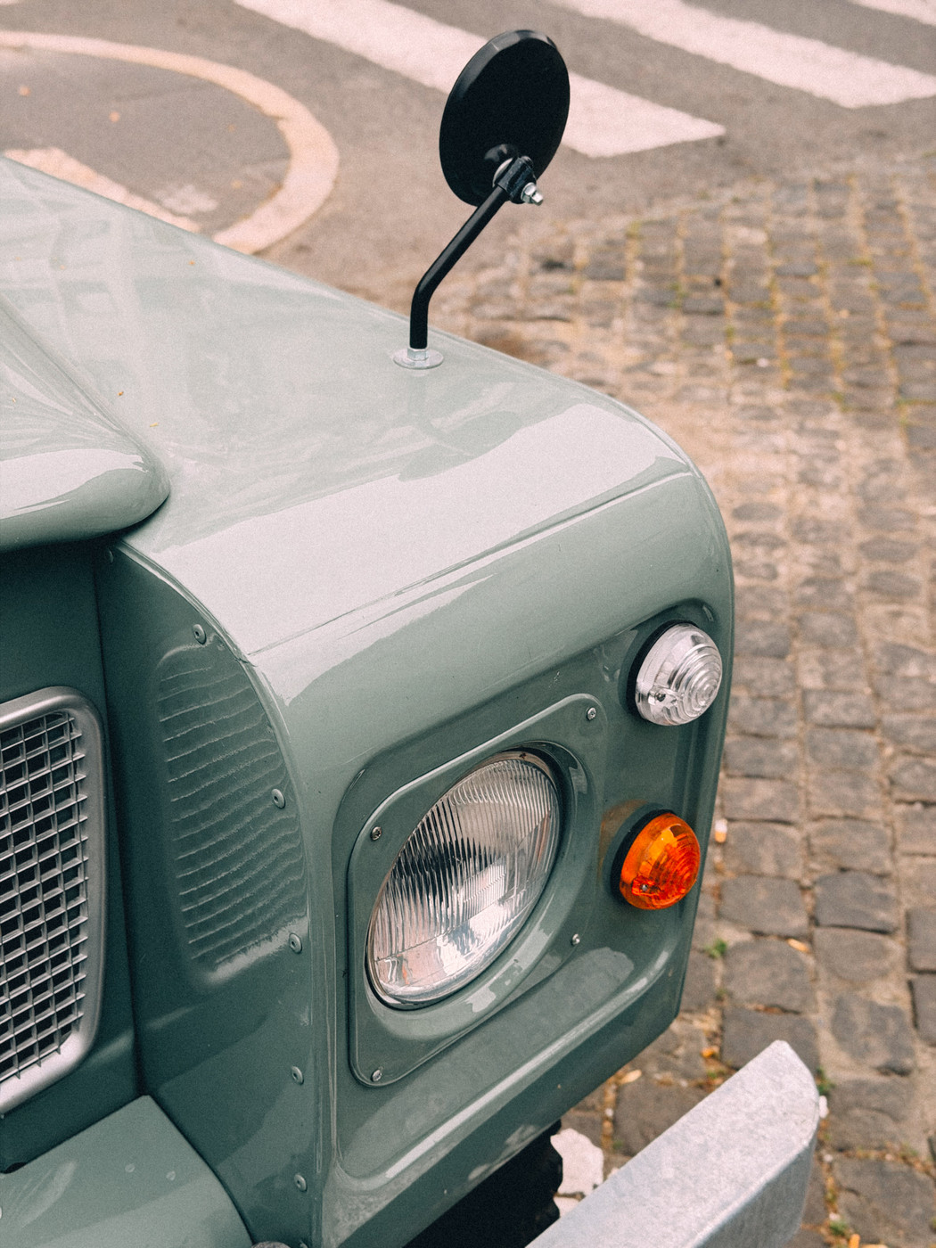 Close-up of the front left corner of a vintage green off-road vehicle, showing the headlight, turn signal, small round side mirror, and part of the grille, parked on a cobblestone street.