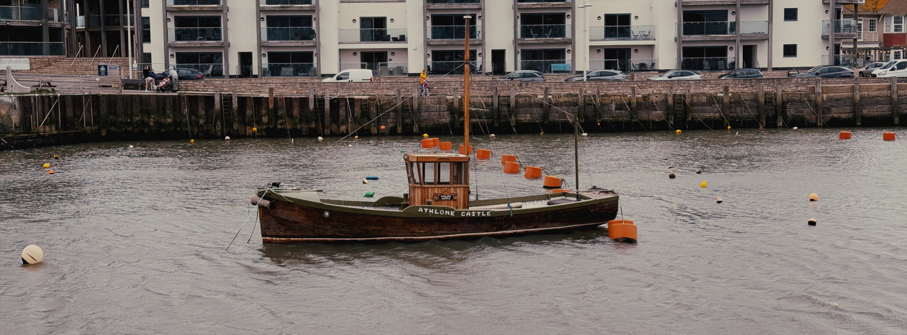 A small wooden boat with a cabin floats on calm water near a dock. The boat is brown with a green deck and has orange buoys attached to it. In the background, there is a row of modern buildings with large windows and parked cars along the waterfront. The scene has a peaceful and quiet mood, with overcast skies reflecting on the water.