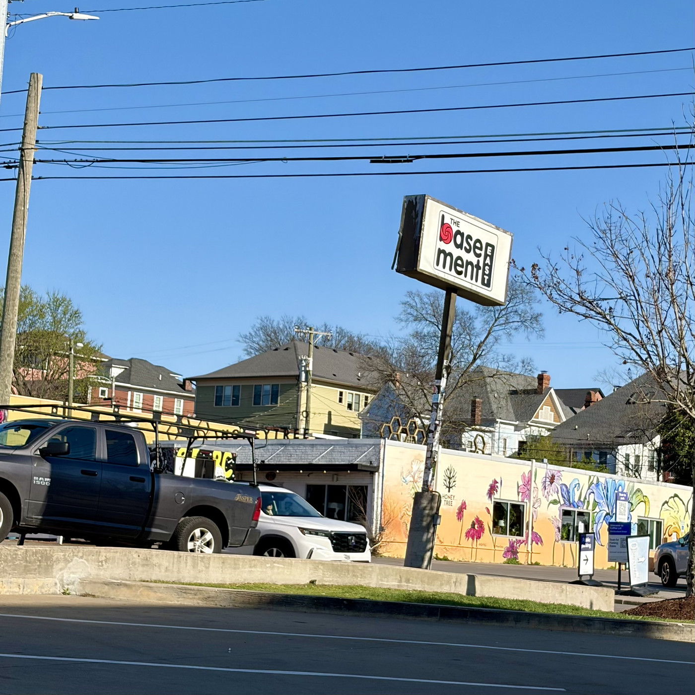 A colorful mural on a building with a sign reading “The Basement” in front of parked cars and houses under a clear blue sky.