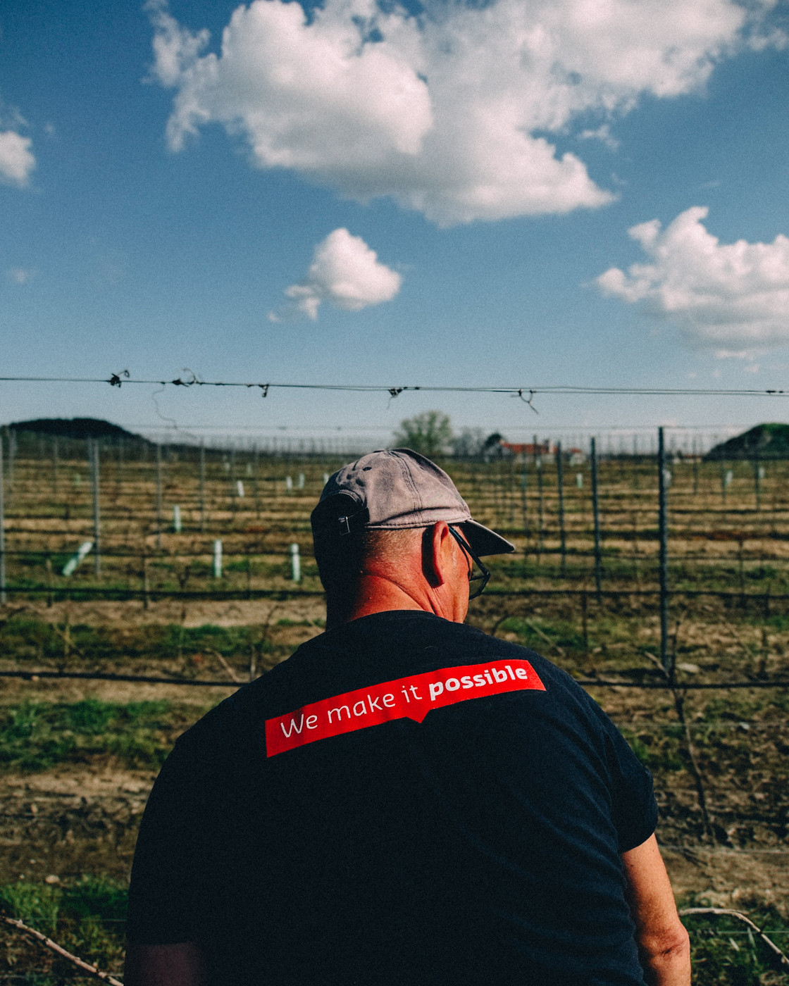 A man wearing a black shirt with the text “We make it possible” and a gray cap stands in a vineyard under a blue sky with scattered clouds.