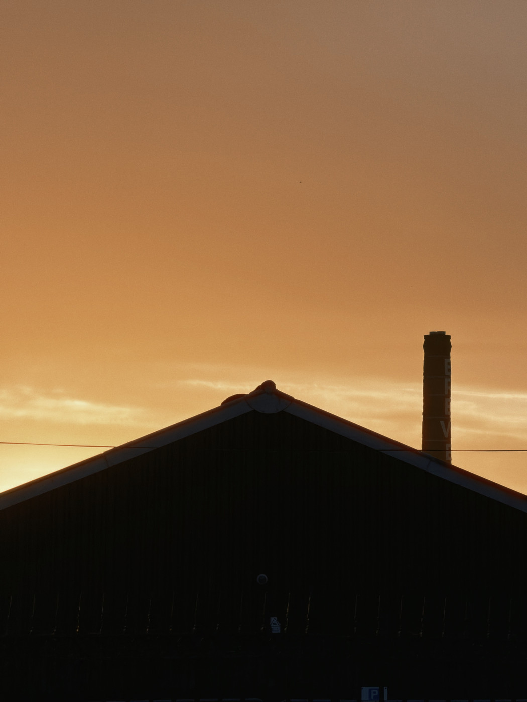 A dark, triangular silhouette of a building roof against a warm, orange and yellow sunset sky, with a tall chimney visible on the right side.