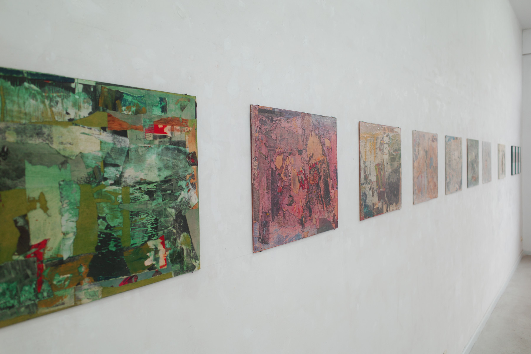 A white gallery wall with a row of abstract square canvases. The nearest piece is a large, multi-colored green-toned collage, followed by several smaller pink, beige, and muted-toned works receding along the wall.