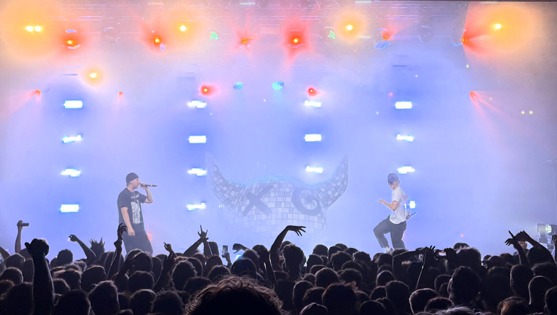 Two male performers on a smoky concert stage. On the left, a man wearing a cap sings into a microphone; on the right, another man in a light shirt is mid-jump. Behind them is a large mosaic horn sculpture, and the stage is lit with bright orange and blue lights while the crowded audience raises their hands in the foreground.