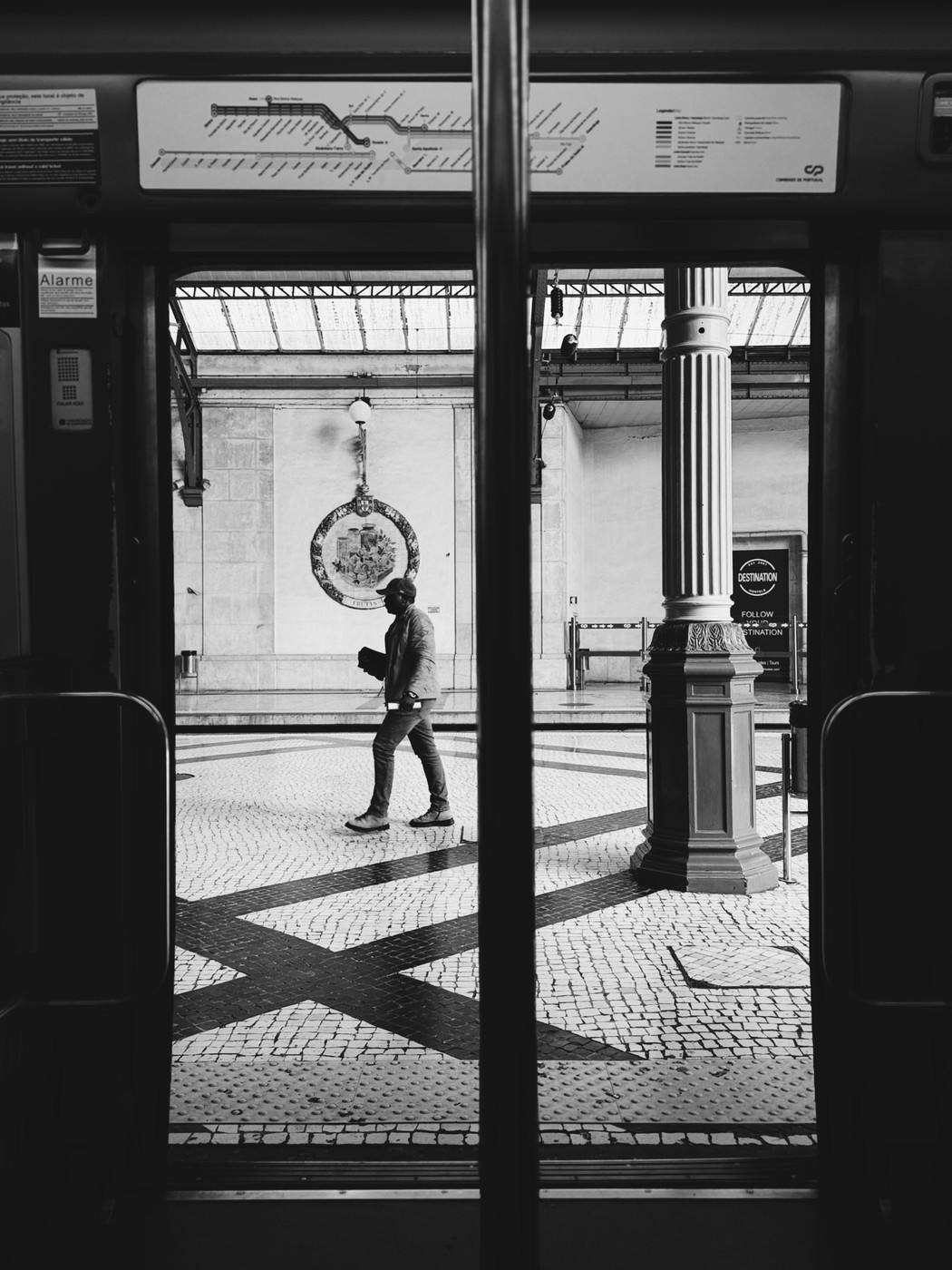 Black and white photo of a man walking across a tiled train station platform, viewed through the open doors of a train with a column and a clock in the background.