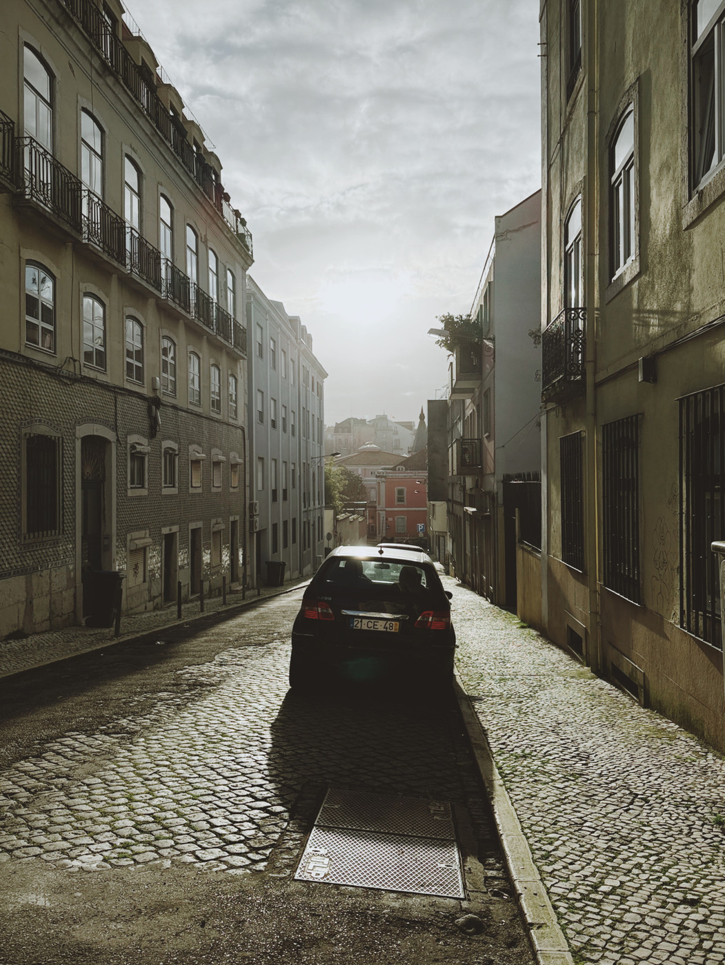 Narrow cobblestone street lined with old buildings and a parked car, with the sun shining through a cloudy sky in the background.