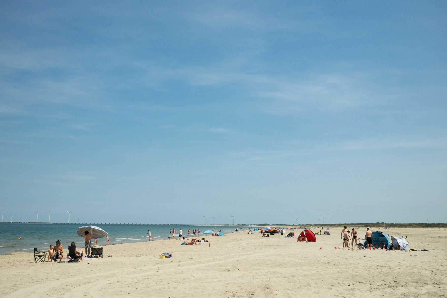 Wide beach scene with people sunbathing and swimming along the shore. Colorful umbrellas and tents dot the sand, while wind turbines line the distant horizon under a bright blue sky.