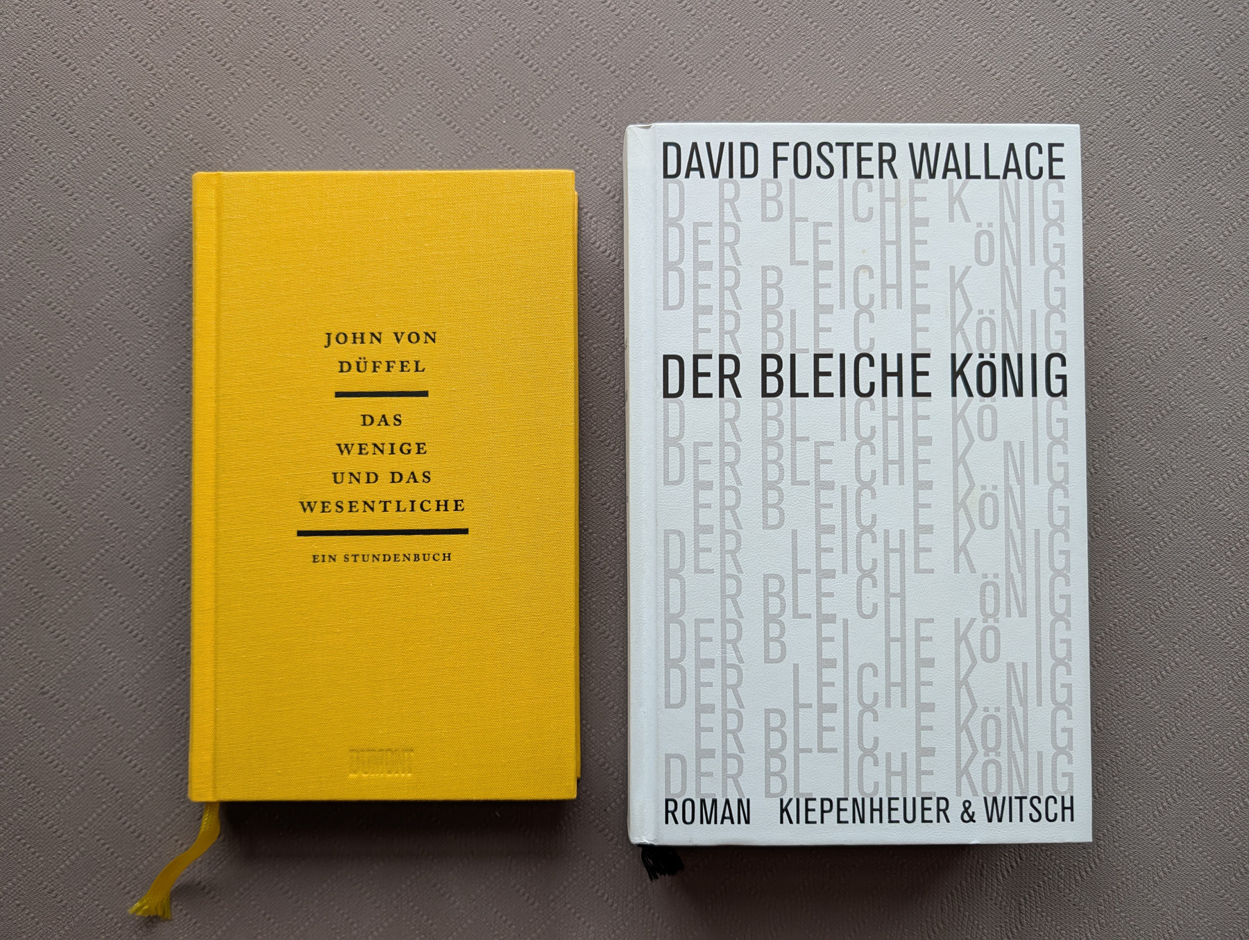 Zwei Bücher liegen nebeneinander auf einem grauen, fein strukturierten Untergrund. Das linke Buch hat einen senfgelben Stoffeinband mit schwarzer Aufschrift: Oben steht “JOHN VON DÜFFEL”, in der Mitte der Titel “DAS WENIGE UND DAS WESENTLICHE”, eingefasst von zwei horizontalen Linien, und darunter der Zusatz “EIN STUNDENBUCH”. Am unteren Rand ist der Verlagsname “DUMONT” leicht eingeprägt. Ein gelbes Lesezeichenband schaut unten aus den Seiten heraus. Das rechte Buch hat ein weißes Cover mit dem schwarzen Autorennamen “DAVID FOSTER WALLACE” am oberen Rand und dem Titel “DER BLEICHE KÖNIG” in der Mitte. Der Hintergrund des weißen Covers ist flächendeckend mit dem Buchtitel in einem sich wiederholenden, hellgrauen und teils unvollständigen Buchstabenmuster bedruckt. Unten auf dem Cover steht “ROMAN KIEPENHEUER & WITSCH”. Unter diesem Buch ragt ein schwarzes Lesezeichenband hervor.