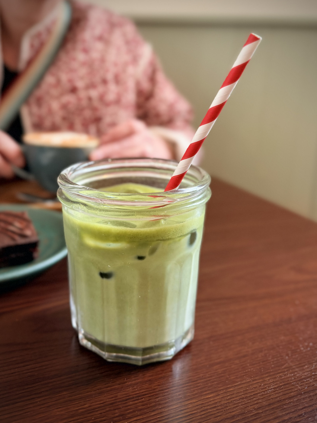 A glass of iced matcha latte with a red-and-white striped straw on a café table, with drinks and cake blurred in the background.