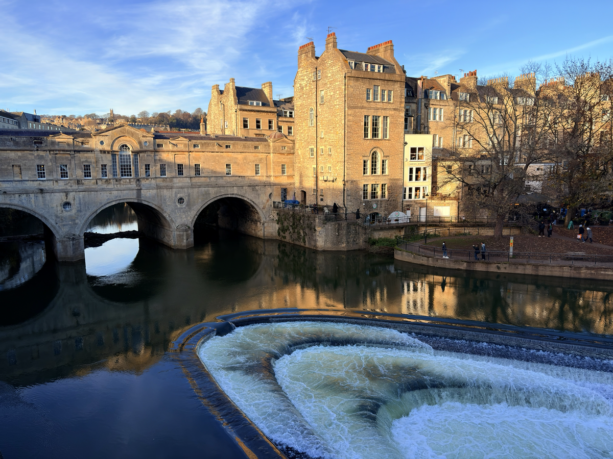 View of the Pulteney Bridge in Bath, England, with its historic stone buildings reflected in the River Avon, and the curved weir in the foreground creating white, cascading water.