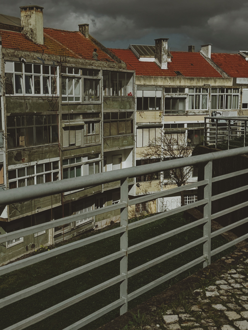 Row of old apartment buildings with weathered facades, red tile roofs, and numerous windows, seen behind a metal railing under a cloudy, dark sky.