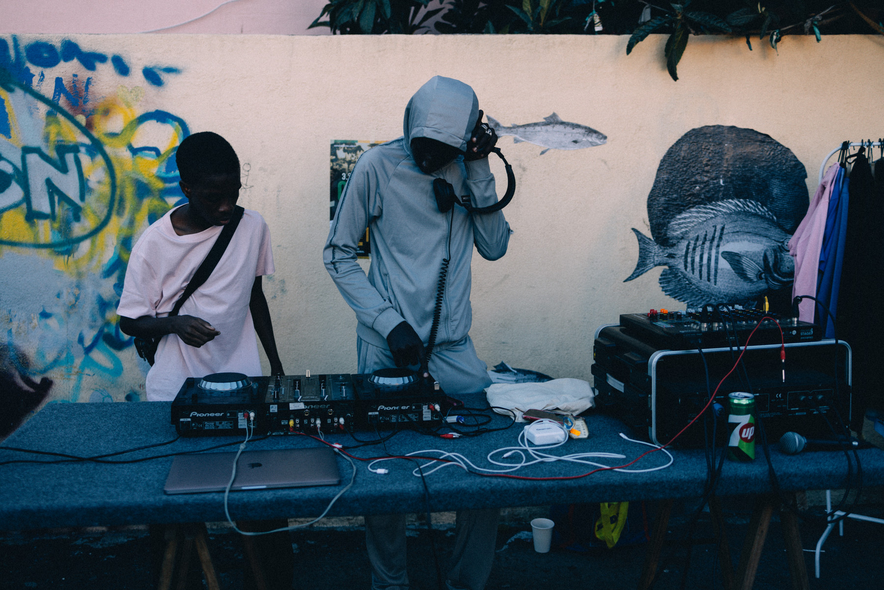 Two people stand at an outdoor DJ setup with turntables and audio equipment against a wall decorated with graffiti and fish murals.