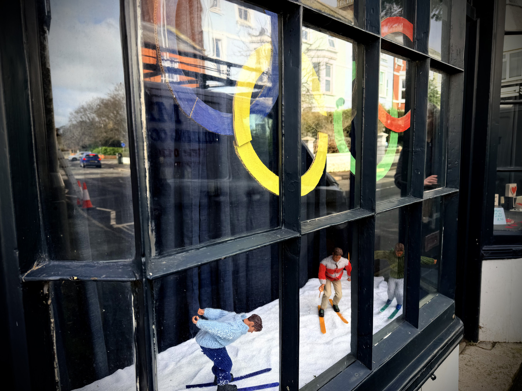 A shop window display with a winter theme. Two small figurines of people ice skating on a snowy surface are positioned in the lower half of the window. Above them, large, colourful paper cut-outs of the Olympic rings hang from the top. The reflection of a street scene, including buildings and trees, is visible in the glass. The overall mood is festive and cheerful, evoking a sense of winter wonder.