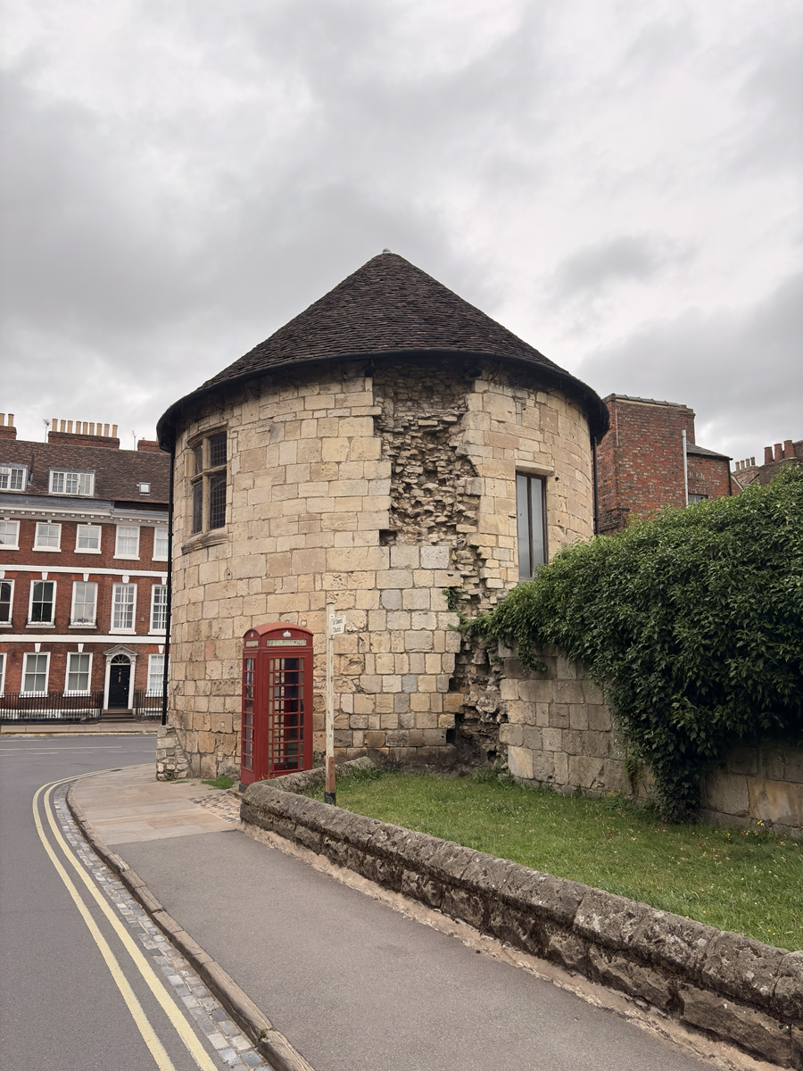 A circular medieval stone tower with a conical tiled roof stands beside a curved street, its weathered masonry showing a large exposed section of rubble. A traditional red British telephone box sits against the tower, with Georgian brick buildings in the background, a low stone wall and grassy verge in the foreground, and an overcast sky overhead.