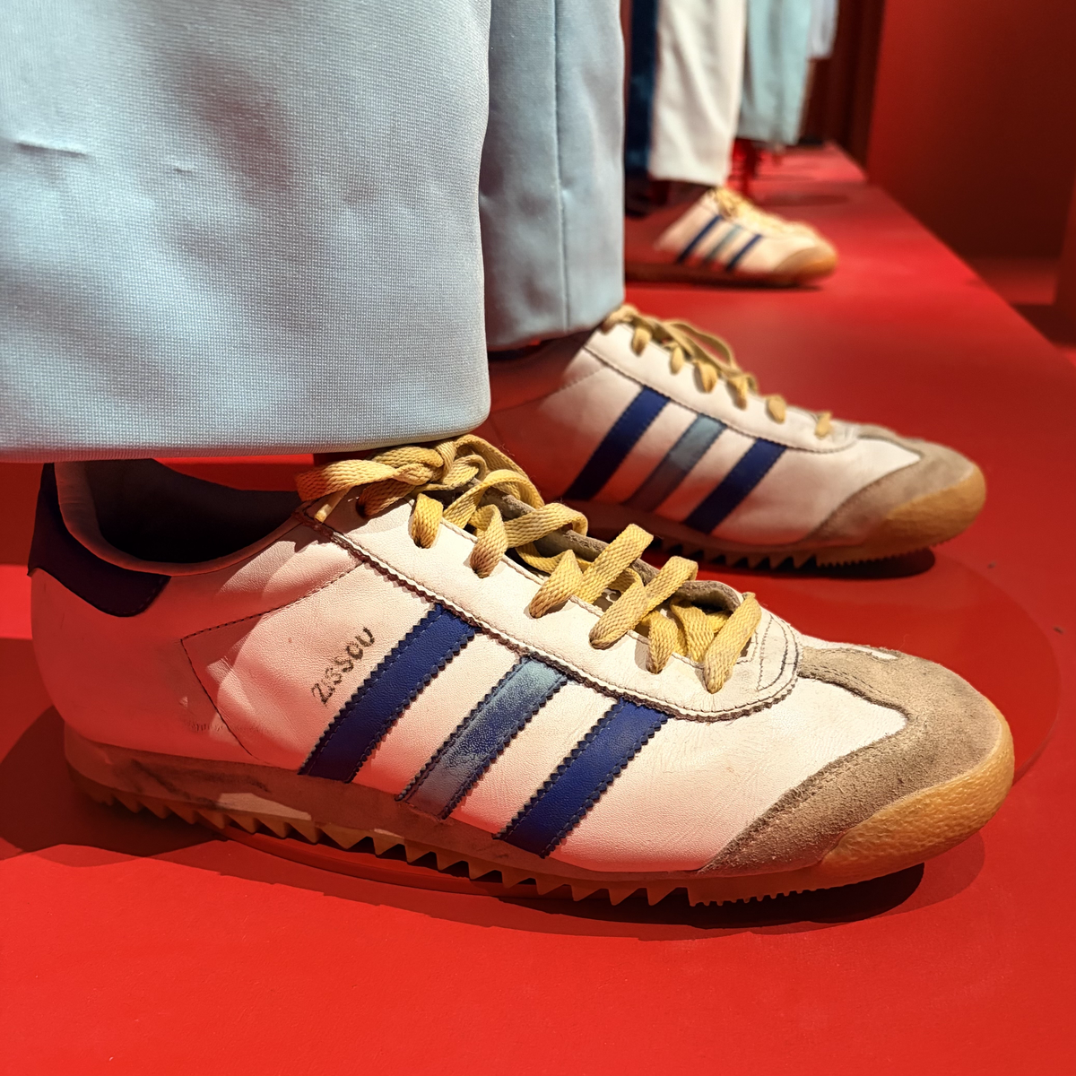 Close-up of a pair of white Adidas trainers with blue stripes and yellow laces, worn with pale trousers, displayed on a bright red surface with another pair visible behind.