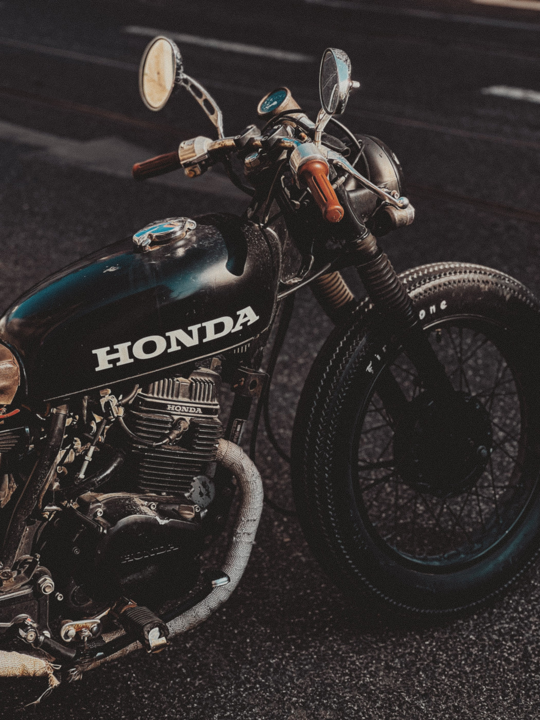Vintage black Honda motorcycle parked on a street, showing the front wheel, handlebars with mirrors, and engine details.