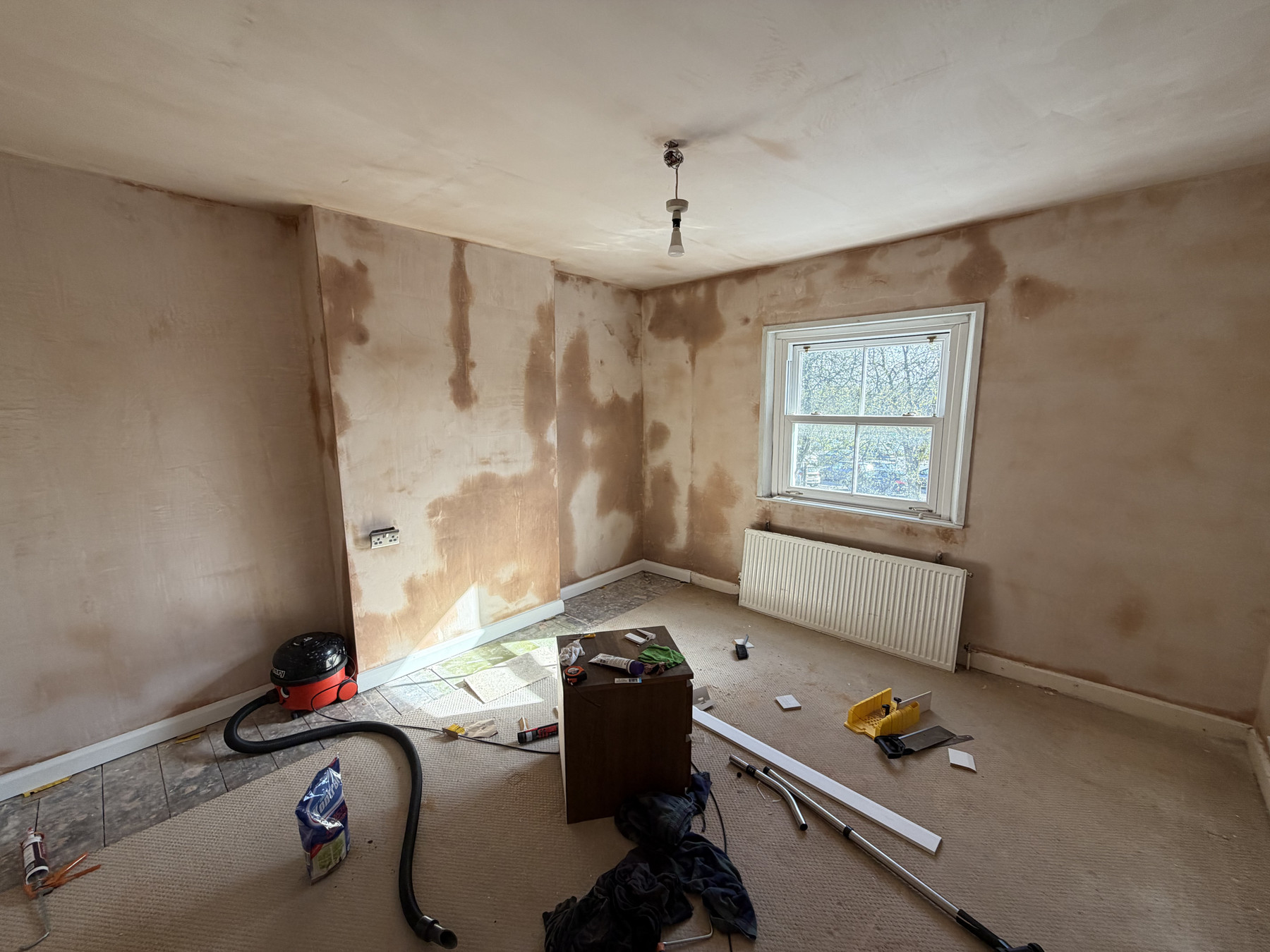 The same room with freshly plastered walls, tools and materials scattered on the floor, and a window with a radiator below.