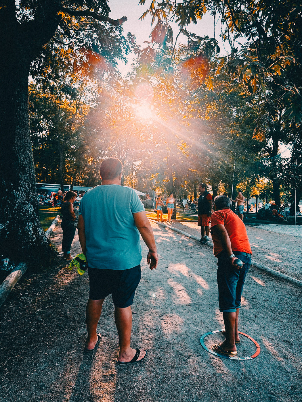 People playing pétanque in a sunlit park, with two men in the foreground, one preparing to throw a ball from a marked circle, surrounded by trees and spectators.