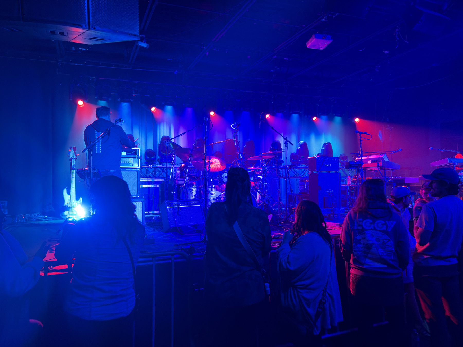 A band performs on stage with colorful lights. Audience stands in front, watching intently. Atmosphere is lively and energetic, typical of a live music concert.