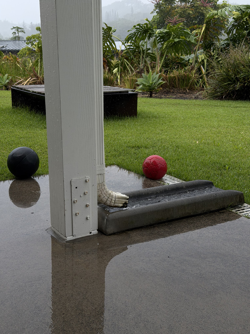 Water pours out of down pipe into a short concrete culvert into a drain. The down pipe is attached to a white wooden post and on one side is a black ball on the other a red ball. Behind that is a lawn with a fire pit on it, and behind that a lush garden bed with trees behind that in the mist.