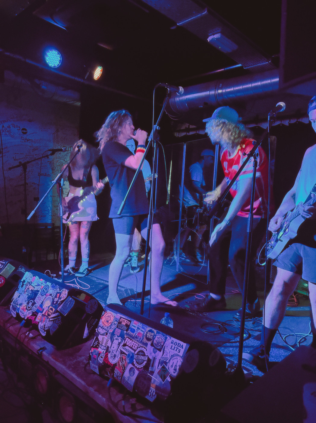 A small club stage with blue/purple lighting shows a live rock band performing. In the foreground center-left, a vocalist with shoulder-length hair sings into a handheld microphone. To the right, a guitarist with long blond hair wearing a red shirt plays. A second guitarist stands to the left, and a drummer is visible behind them. The edge of the stage is covered with stickers and flyers, and a graffiti-covered wall and audience silhouettes are in the dark background.