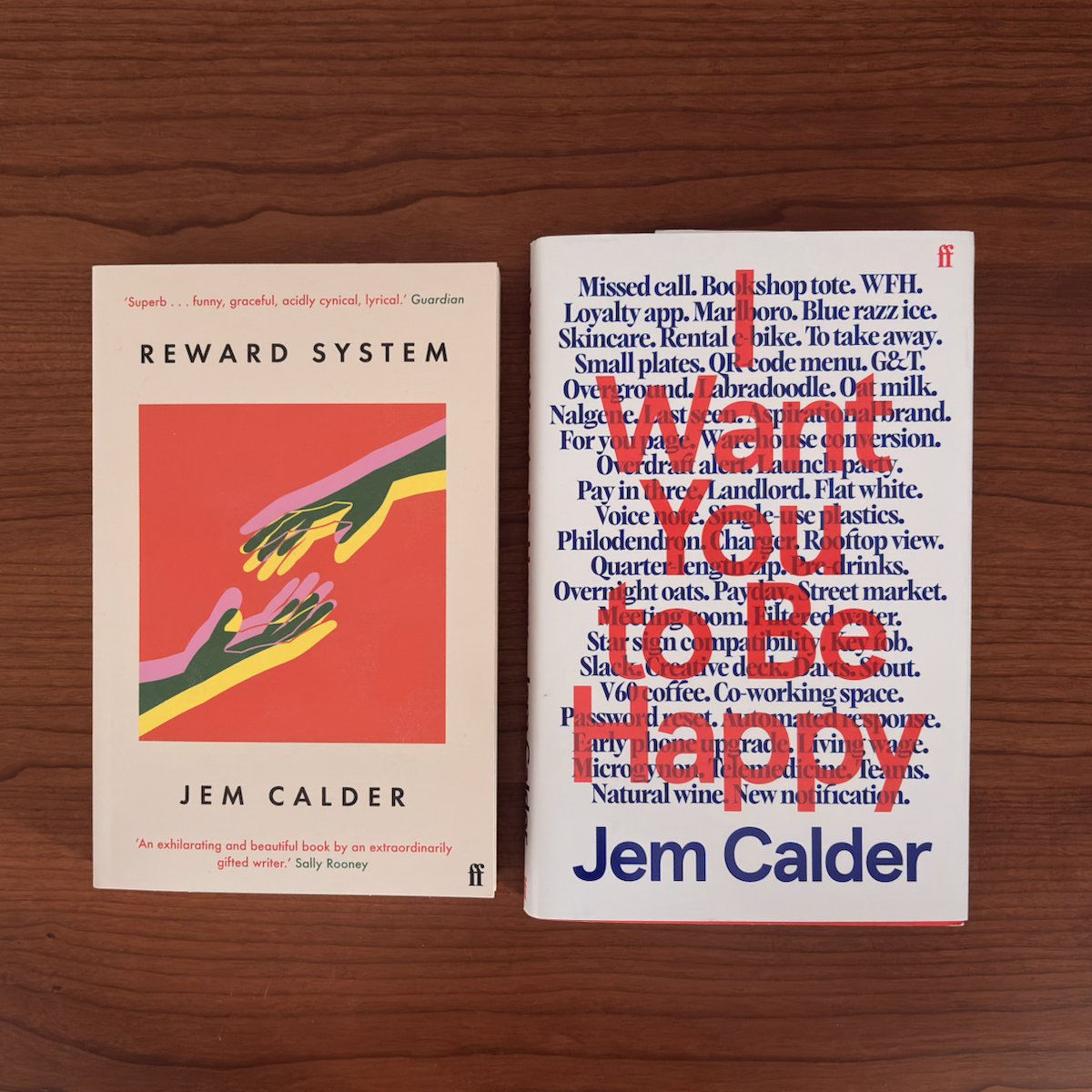 Two books by Jem Calder are placed side-by-side on a wooden surface. The book on the left has a cream cover with a red square in the center, featuring a graphic of two hands reaching toward each other, and is titled ‘Reward System’. The book on the right has a white cover with a list of modern, everyday phrases printed in blue, and the title ‘I Want You to be Happy’ overlaid in large, translucent red text.