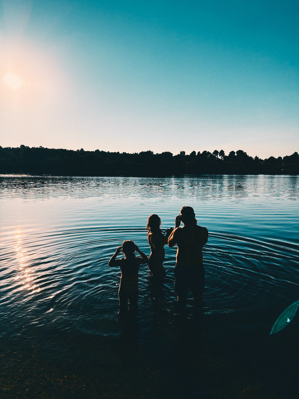 Silhouettes of three people standing in shallow water of a calm lake at sunset with trees in the background.