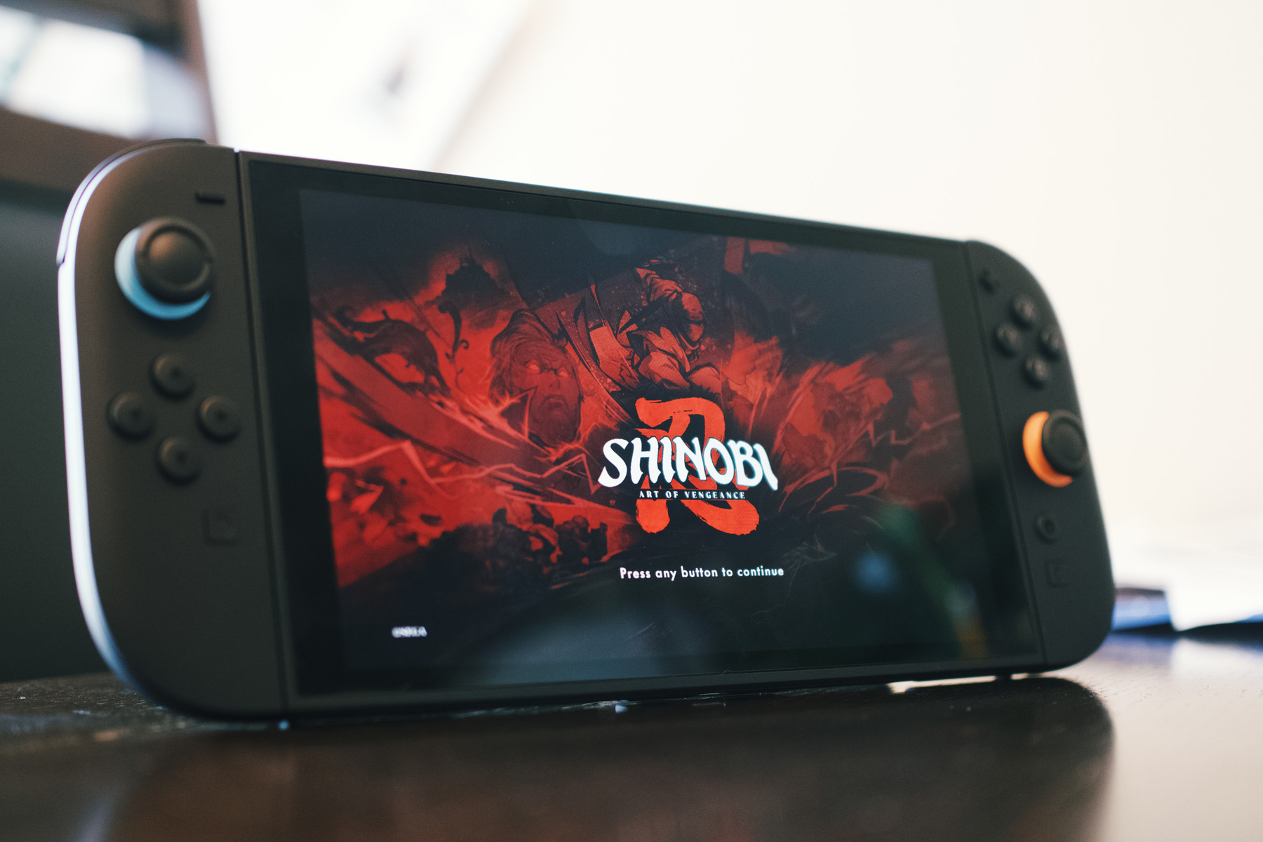 A handheld gaming console displaying the title screen for Shinobi: Art of Vengeance. The screen features a dramatic red and black illustration with ninja figures and swords in the background, and the game's logo centered in white and red text. A prompt at the bottom reads, "Press any button to continue." The console has black Joy-Cons with blue and orange joystick accents.