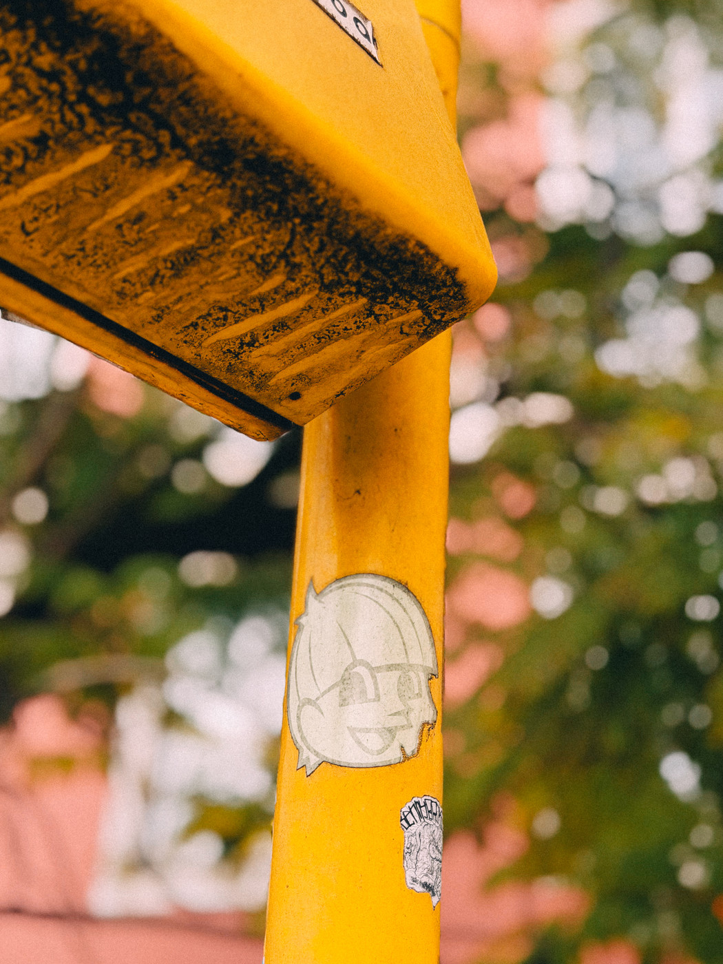 Close-up of a yellow metal pole with a worn cartoon face sticker, with blurred green trees and a pink building in the background.