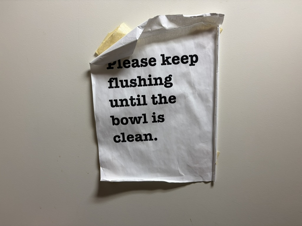 A ragged paper taped to a wall that says, "Please keep flushing until the bowl is clean."