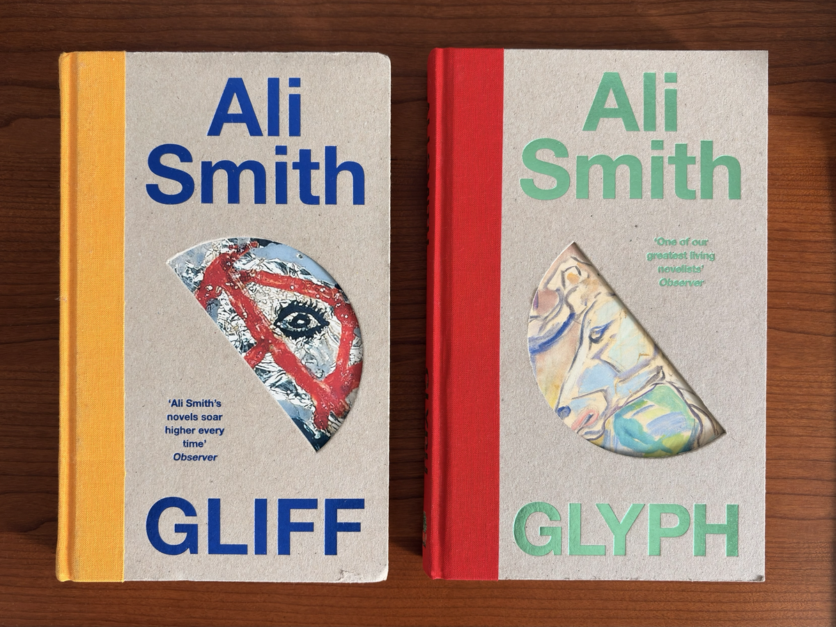 Two hardback Ali Smith novels, *Gliff* and *Glyph*, lying side by side on a wooden surface. *Gliff* has a mustard-yellow cloth spine, bold blue title and author text, and a semicircular cut-out revealing part of an eye beneath red painted lines. *Glyph* has a bright red cloth spine, pale green title and author text, and a matching cut-out revealing an abstract pastel illustration. Both covers are cream-coloured with a similar minimalist design.