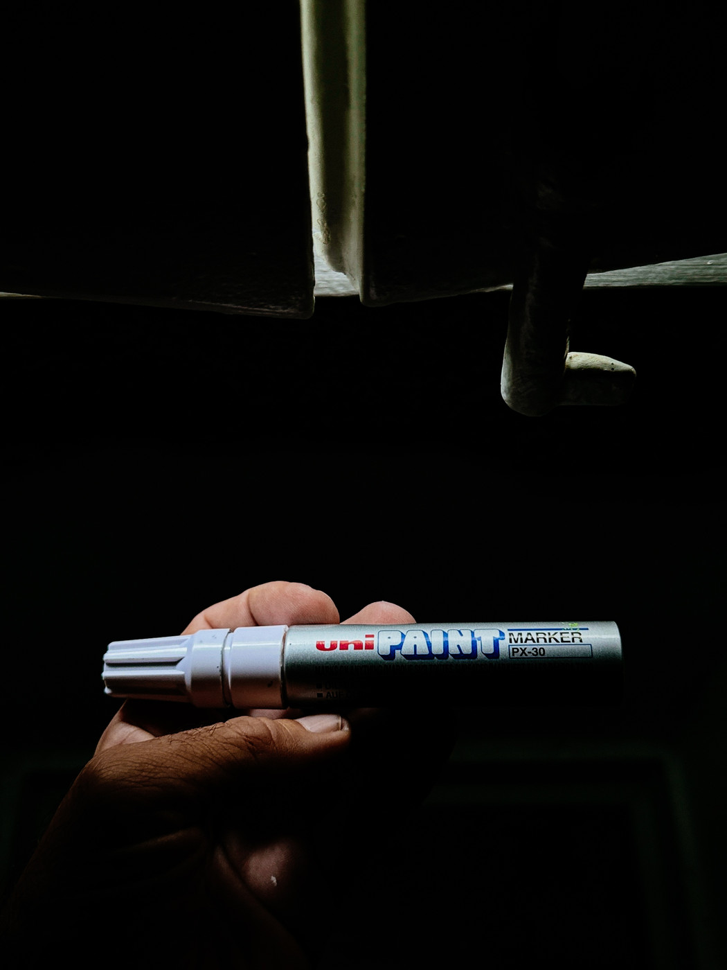 A hand holding a silver Uni Paint Marker PX-30 with a white cap against a dark background with a small amount of light shining from above.