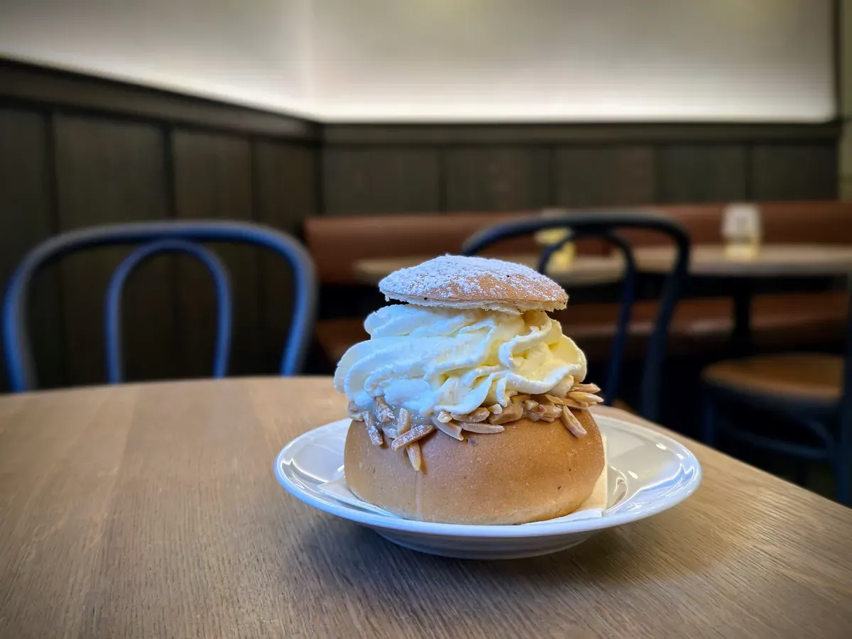 A Swedish semla on a white plate on a wooden café table, a cardamom bun filled with whipped cream and slivered almonds, topped with a lid dusted with powdered sugar.