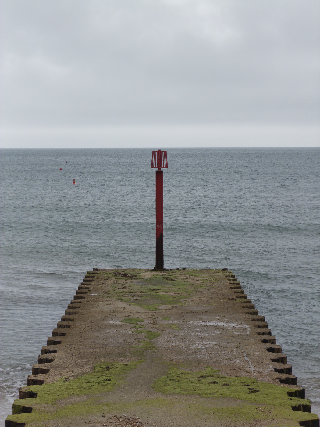 Red metal navigation marker atop a concrete groyne covered in green algae, extending into a grey sea under an overcast sky.