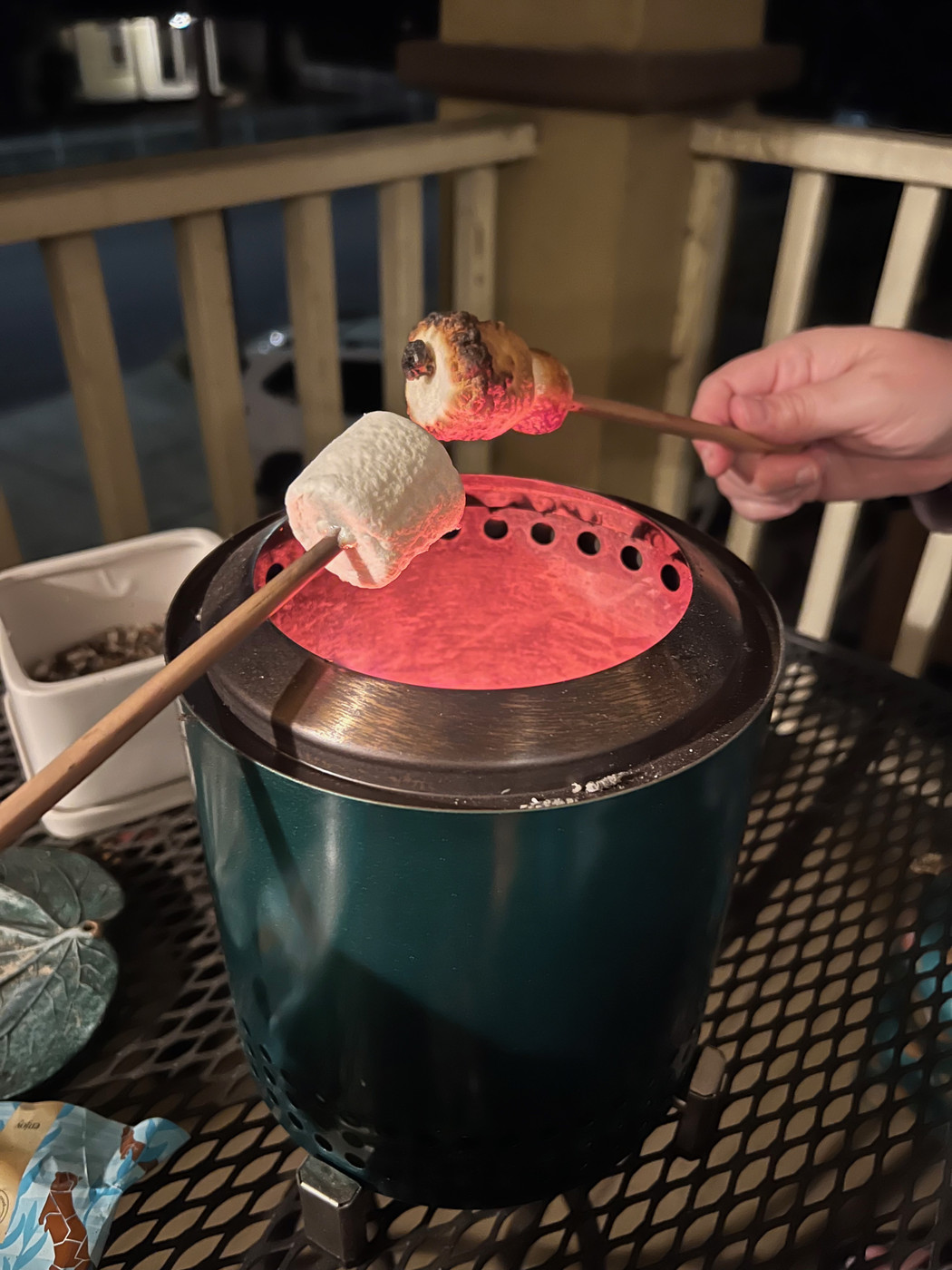 A small green fire pit on a porch table, with two people holding marshmallows over the glowing orange top, speared onto chopsticks.
