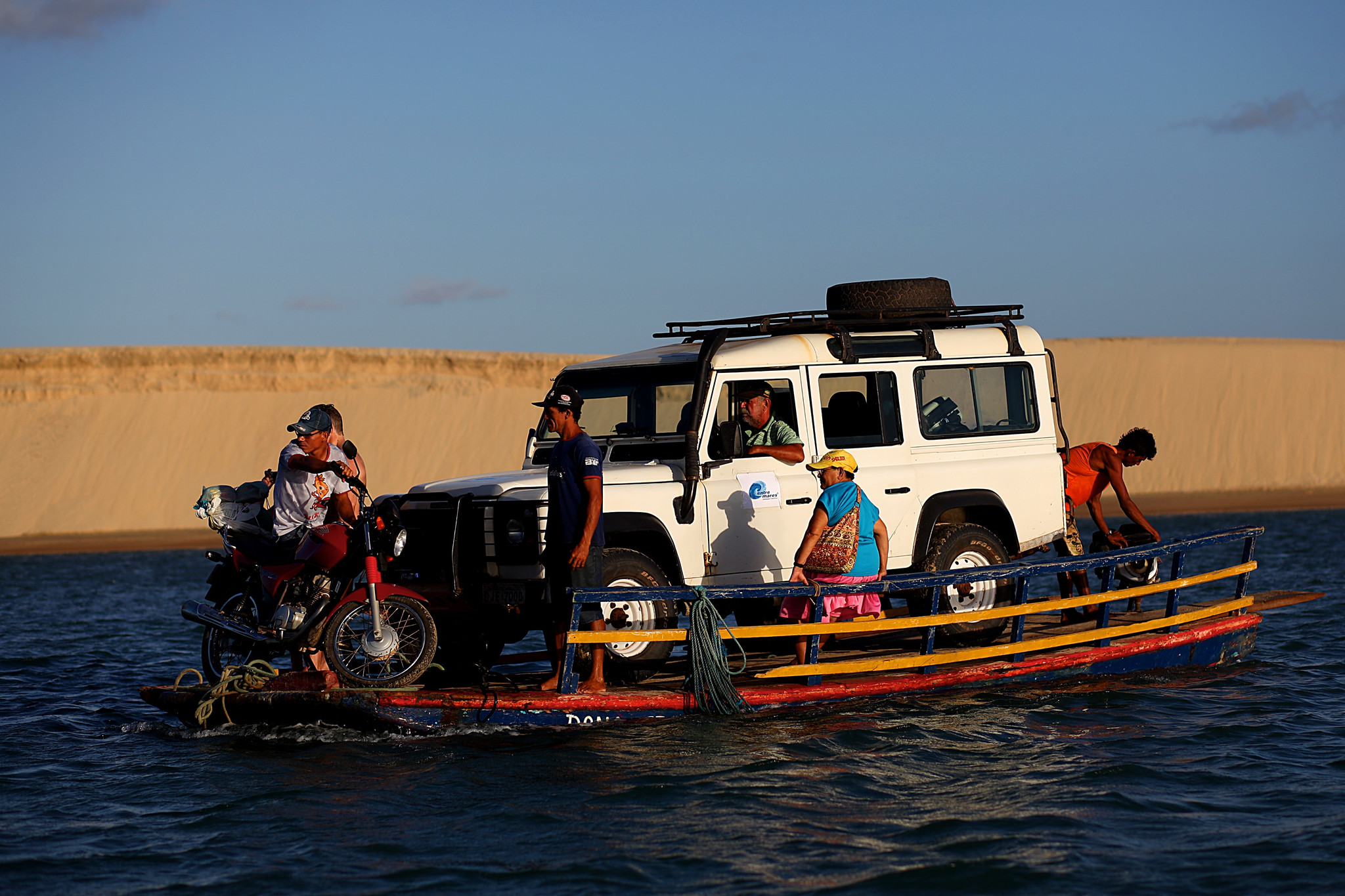 A small wooden ferry carrying a white Land Rover Defender, a motorcycle, and several people across a body of water with sand dunes in the background.