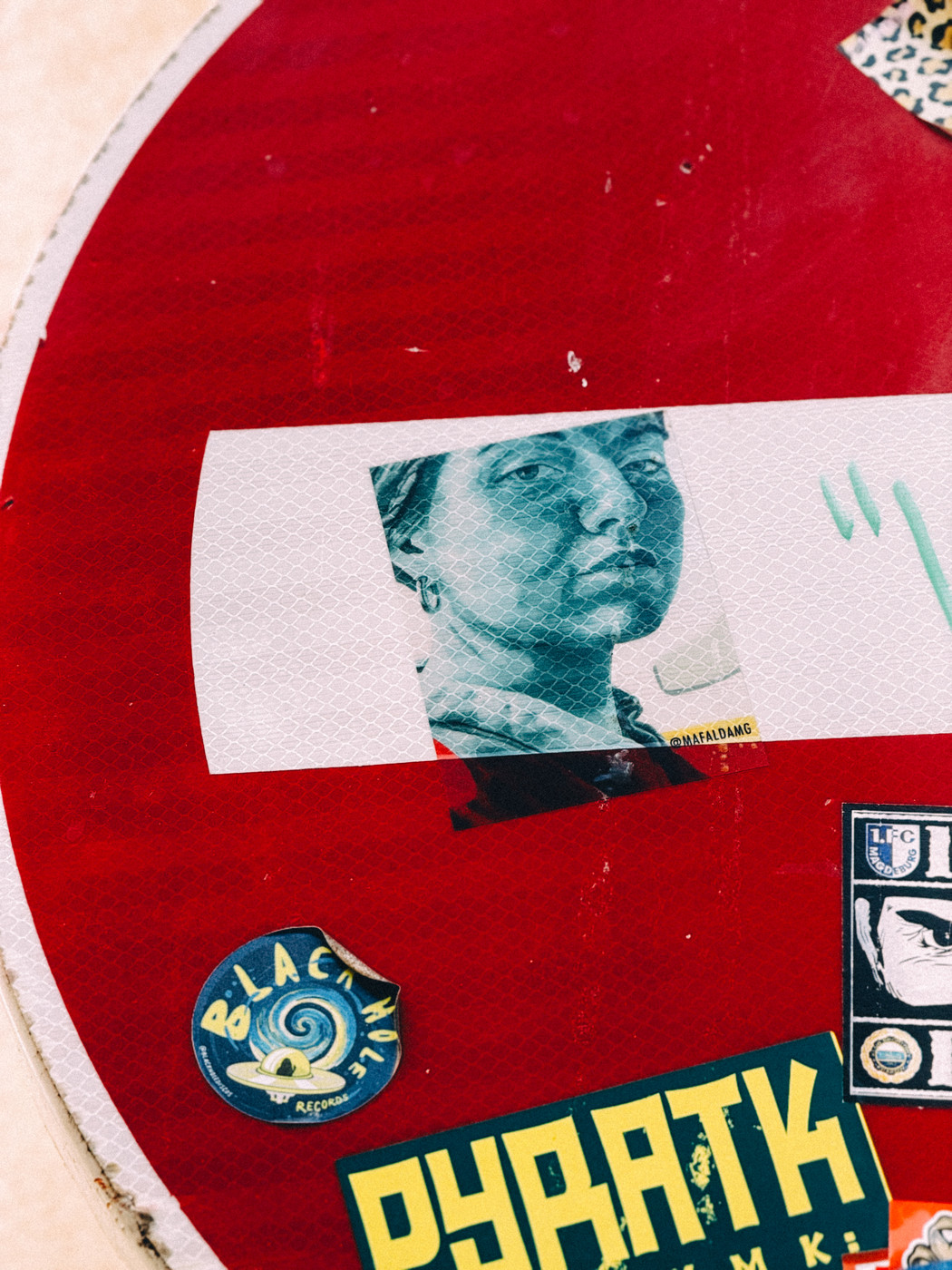Close-up of a red street sign covered with various stickers, including a teal portrait of a woman, a “Black Hole Records” logo, and a yellow and green “PYRATK” sticker.