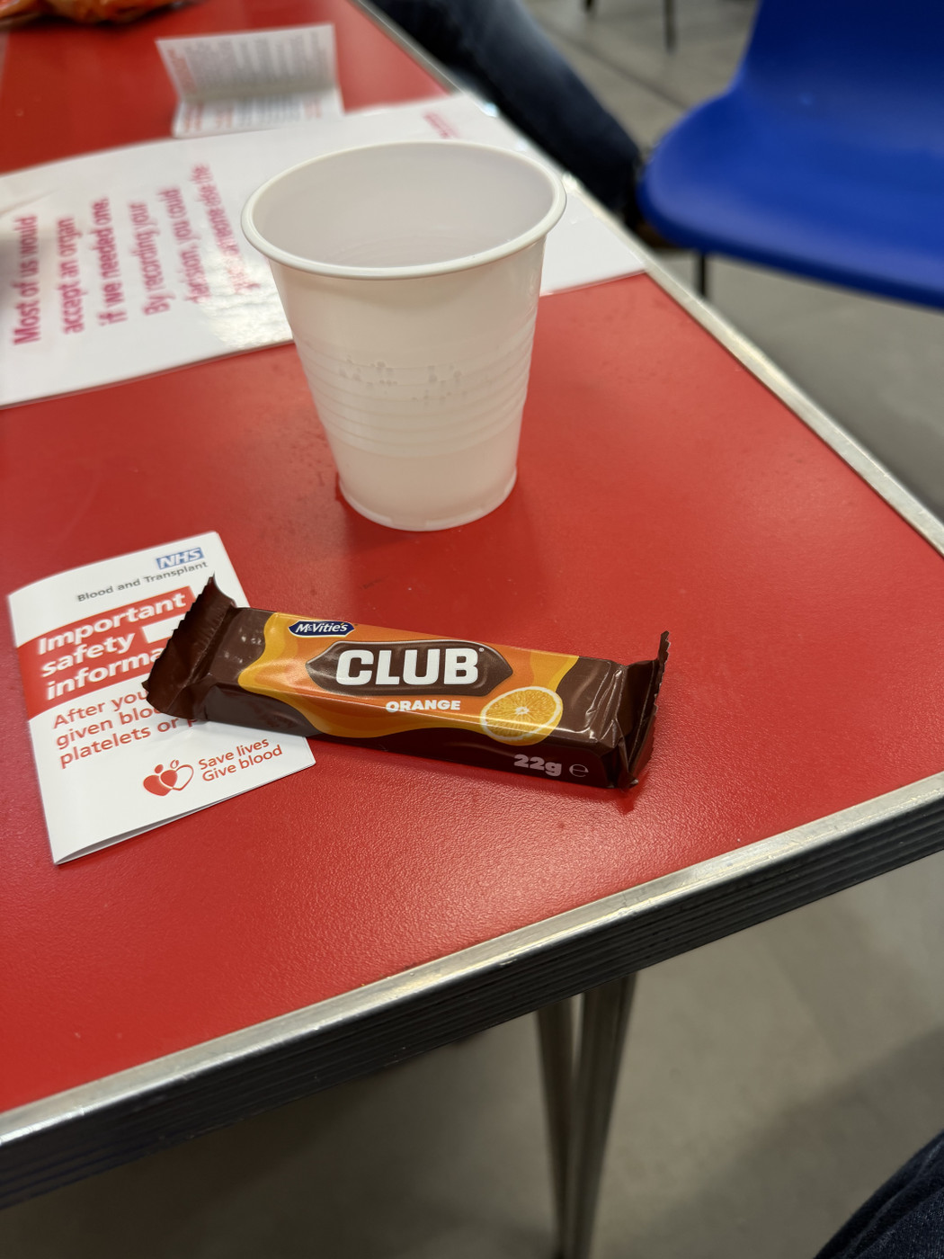A red table with a white plastic cup, a McVitie’s Club Orange biscuit bar, and an NHS Blood and Transplant leaflet titled “Important safety information after you have given blood, platelets or plasma.”