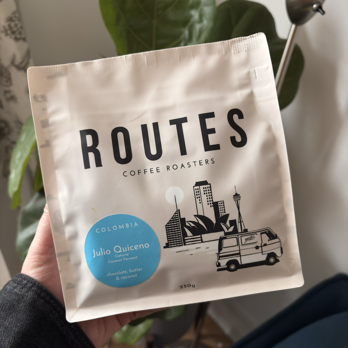 A hand holding a matte white bag of coffee labeled 'Routes Coffee Roasters', with a blue sticker reading 'Colombia, Julio Quiceno, Caturra, Coconut Ferment' and tasting notes of chocolate, butter, and coconut, illustrated with a city skyline and van.