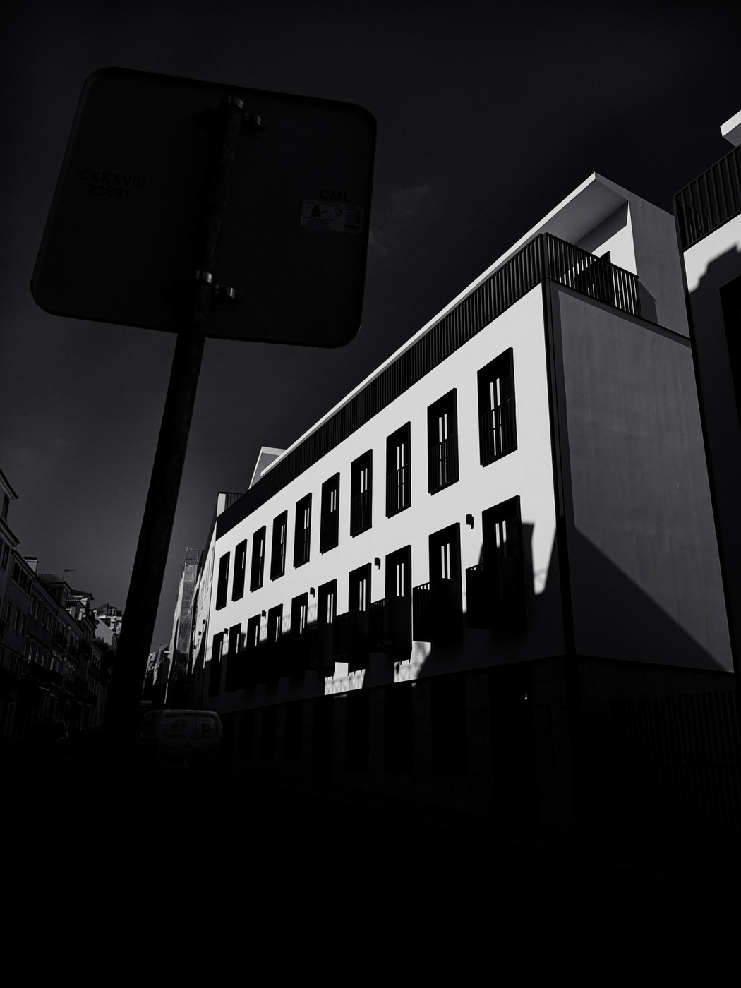 Black and white photo of a modern multi-story building with rectangular windows, deep shadows, and a silhouetted street sign in the foreground.