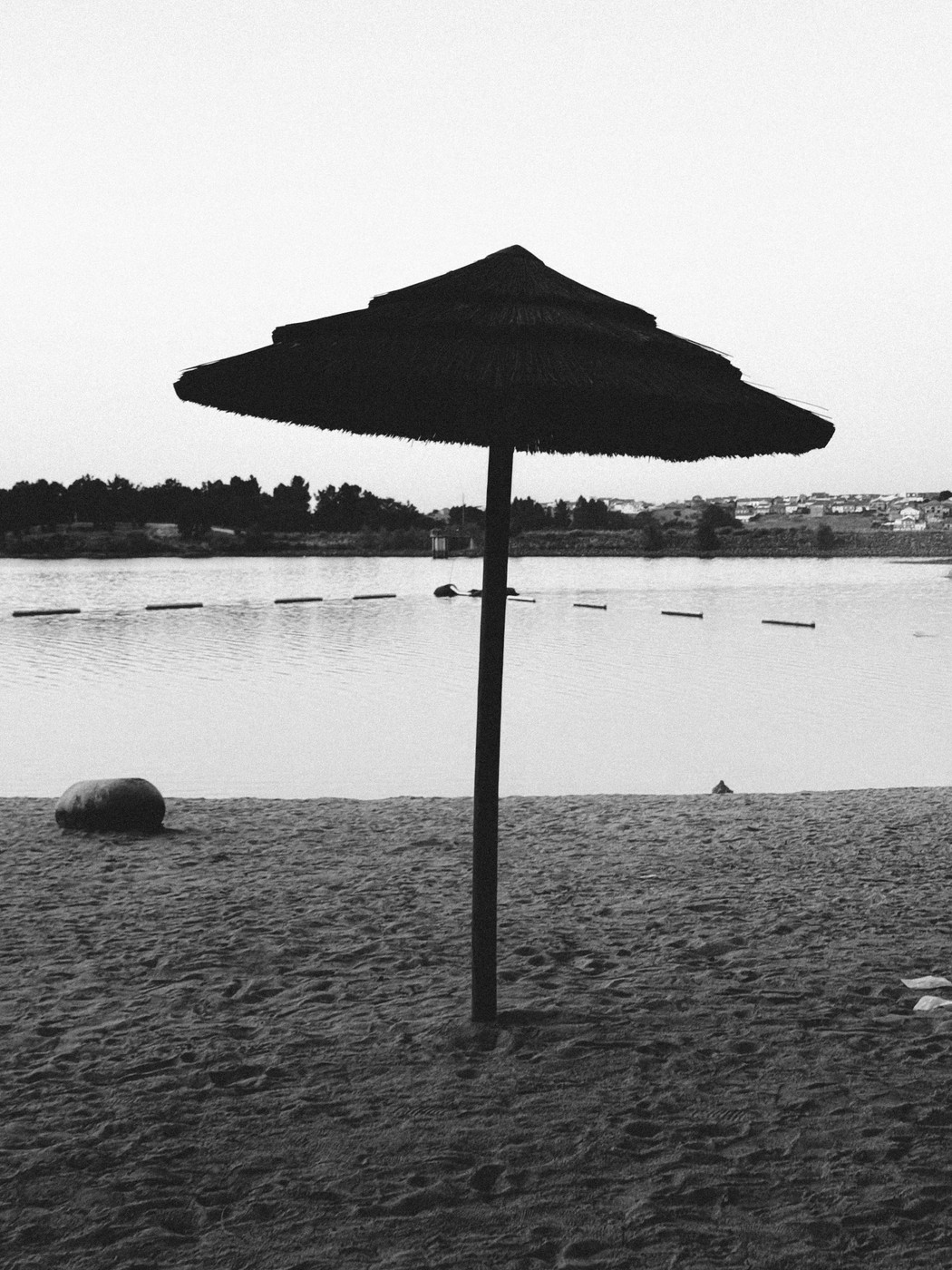 Silhouette of a thatched beach umbrella on a sandy shore by a calm lake with distant trees and houses under a clear sky.