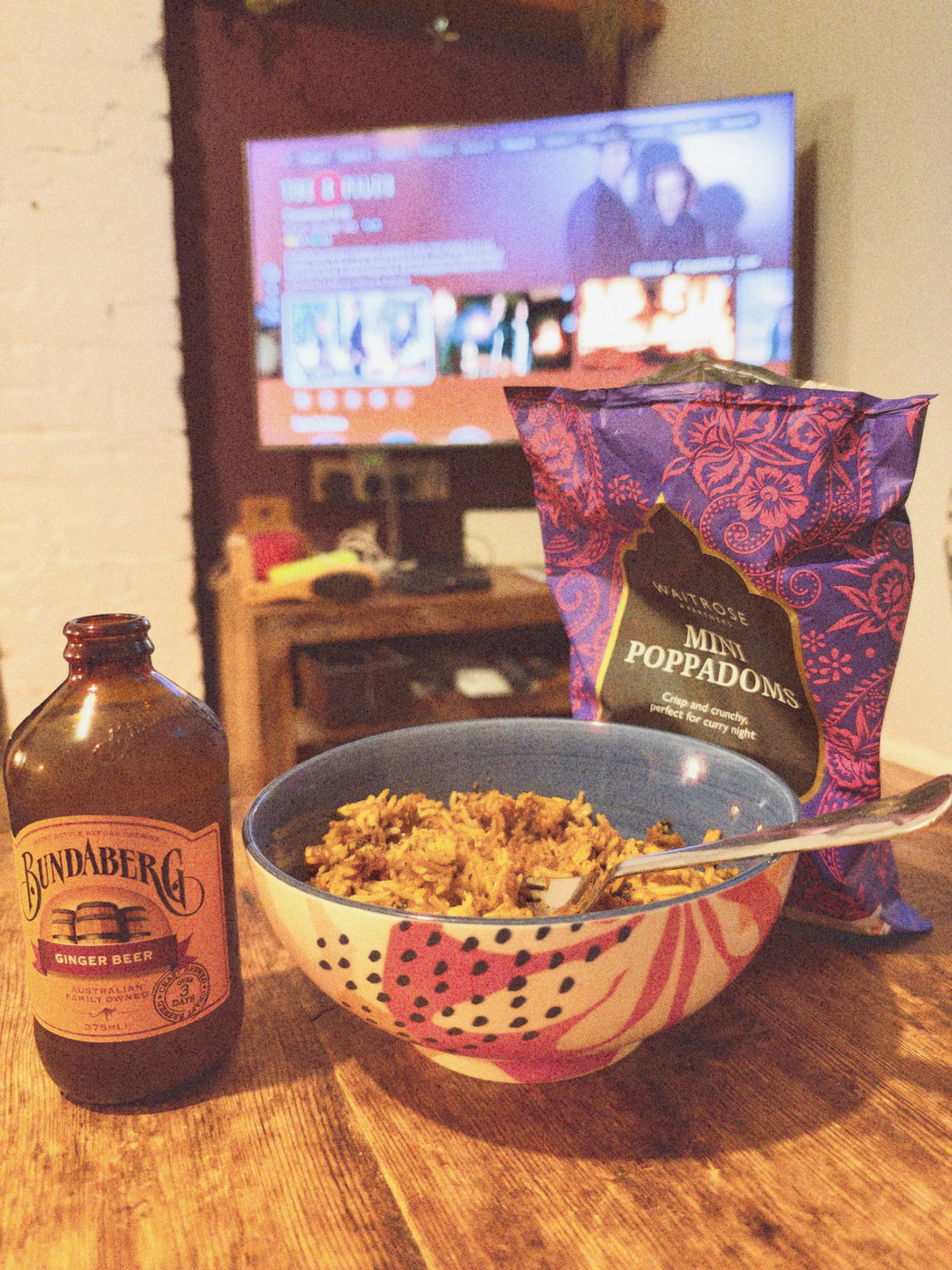 Bowl of curried rice with a fork, alongside a bottle of Bundaberg ginger beer and a bag of mini poppadoms, TV streaming menu blurred in background.