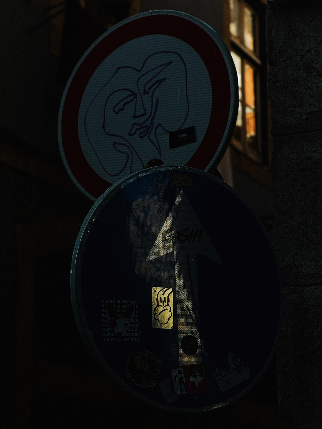 Two overlapping street signs, one with a red border and a face doodle, the other a blue one-way sign covered with various stickers.