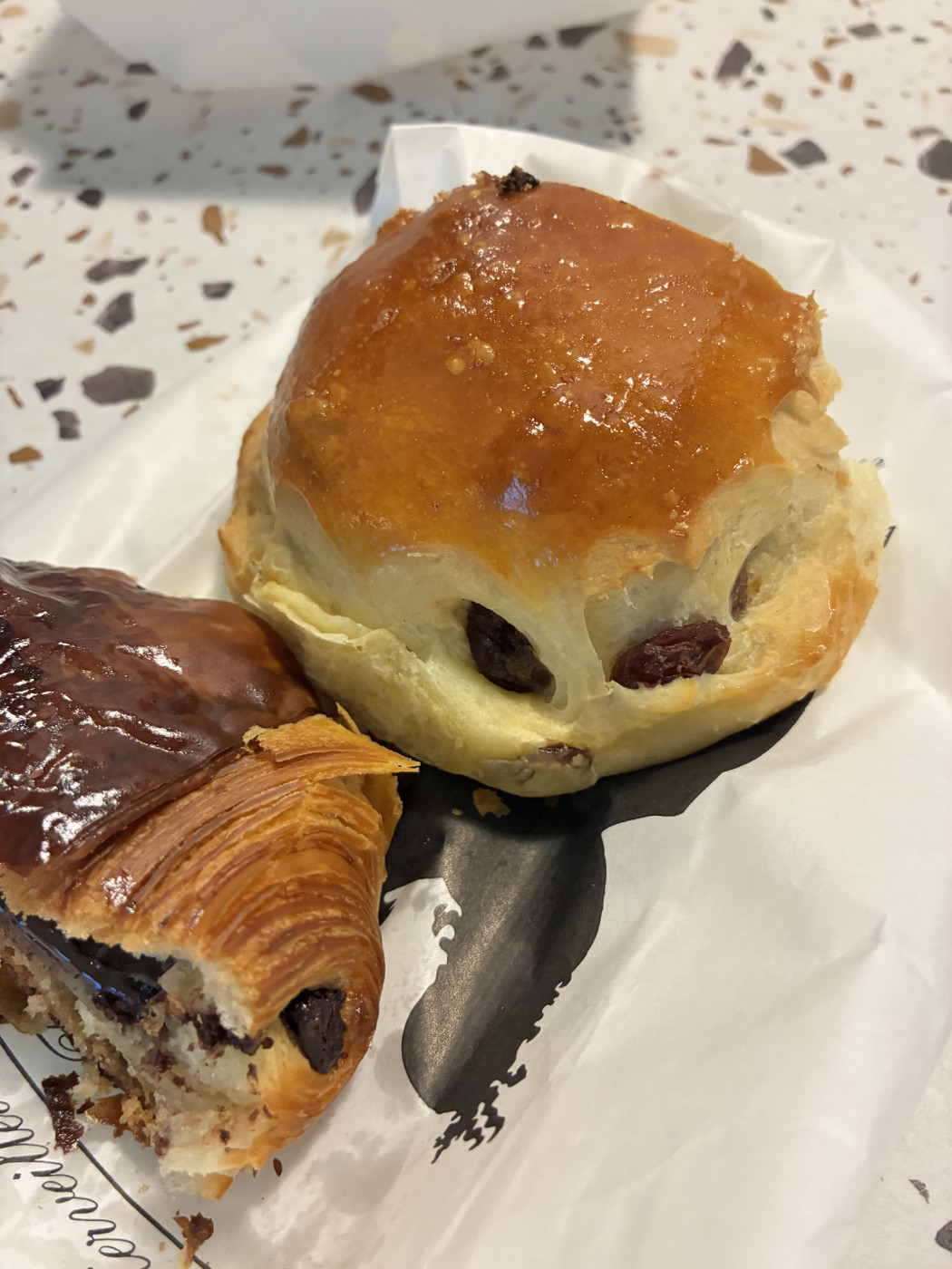 Amber-glazed raisin brioche bun beside a chocolate pastry with flaky layers, on white bakery paper with black logo.