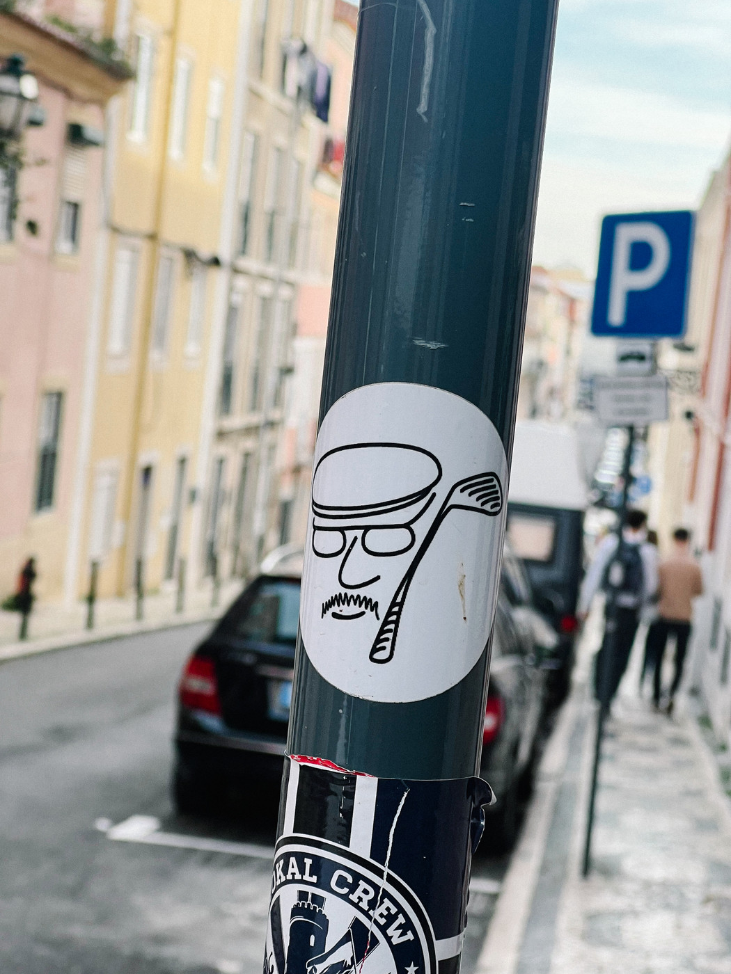 Sticker of a cartoon face wearing glasses, with a golf club, on a street pole in an urban neighborhood with cars parked and people walking in the background.